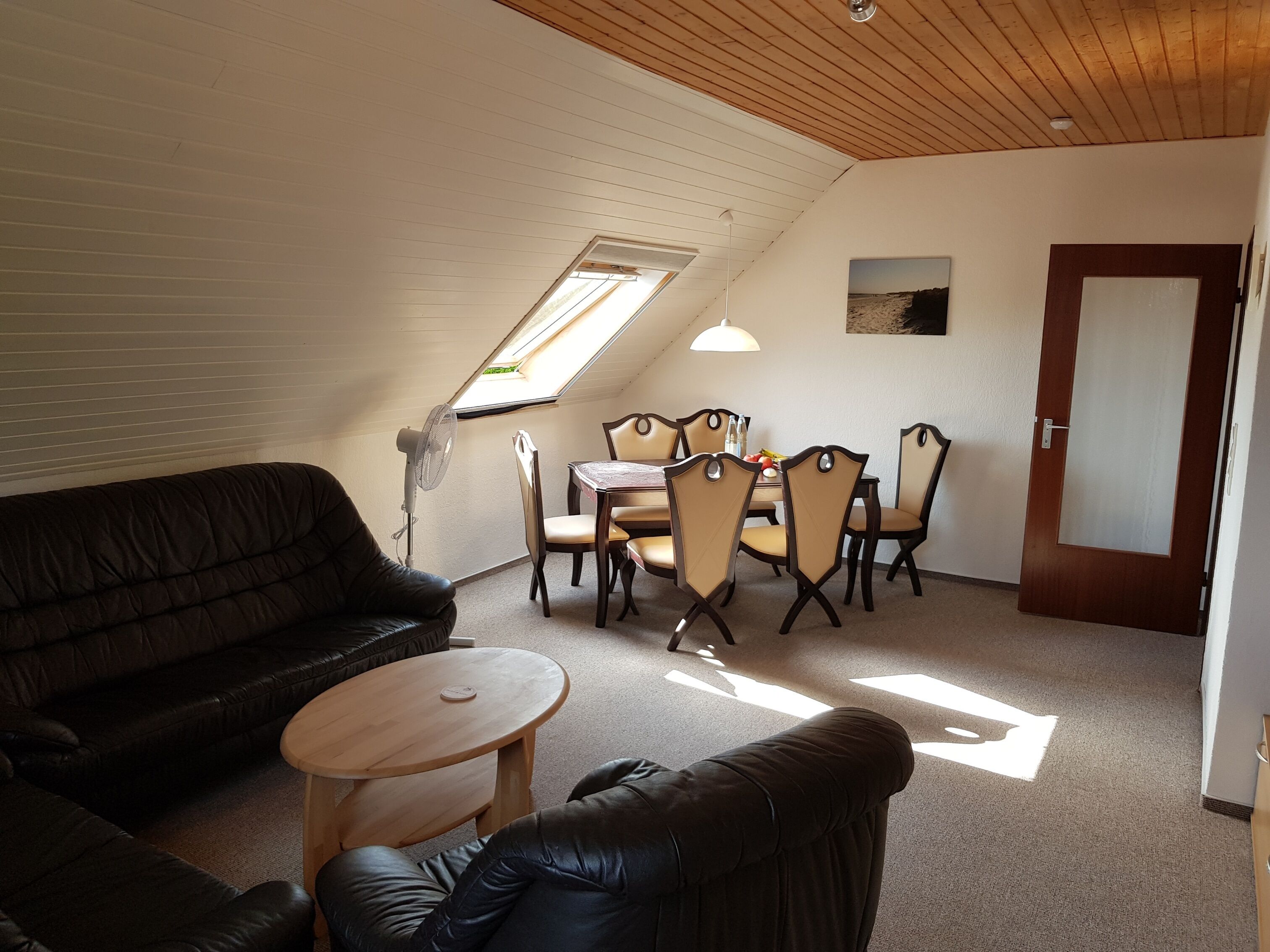 A cozy room with a sloped ceiling, a wooden ceiling, a dining table with chairs, a sofa, and a small round table.