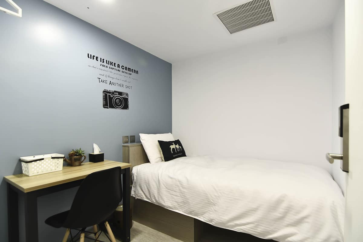 Ein ordentlich eingerichtetes Schlafzimmer mit Bett, Schreibtisch und Stuhl. An der Wand befindet sich ein motivierendes Zitat und eine Illustration einer Kamera.