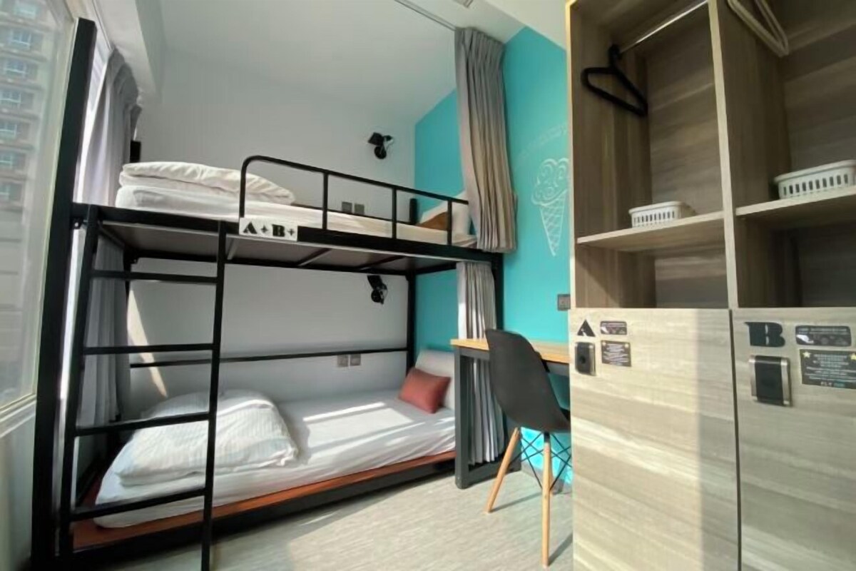A bunk bed room with a desk and chair, a window with curtains, and a wardrobe with hanging items.