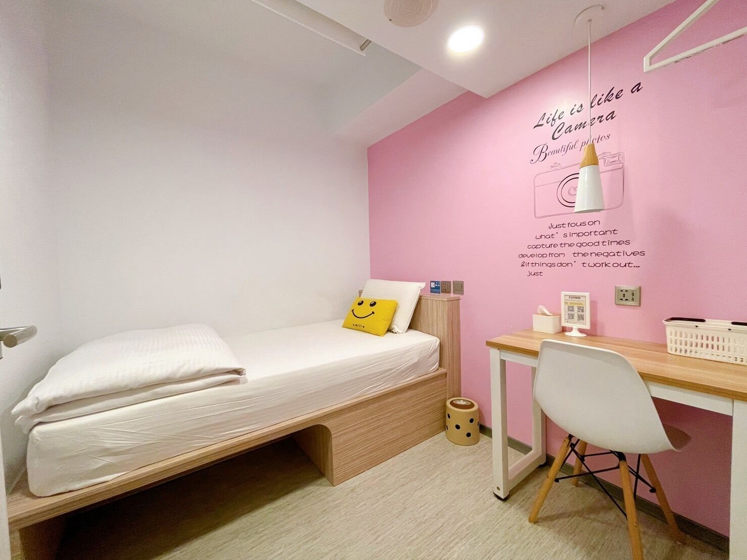 Una habitación pequeña y ordenada con una cama, un escritorio y una silla. Las paredes son rosas y hay una frase motivadora en la pared.