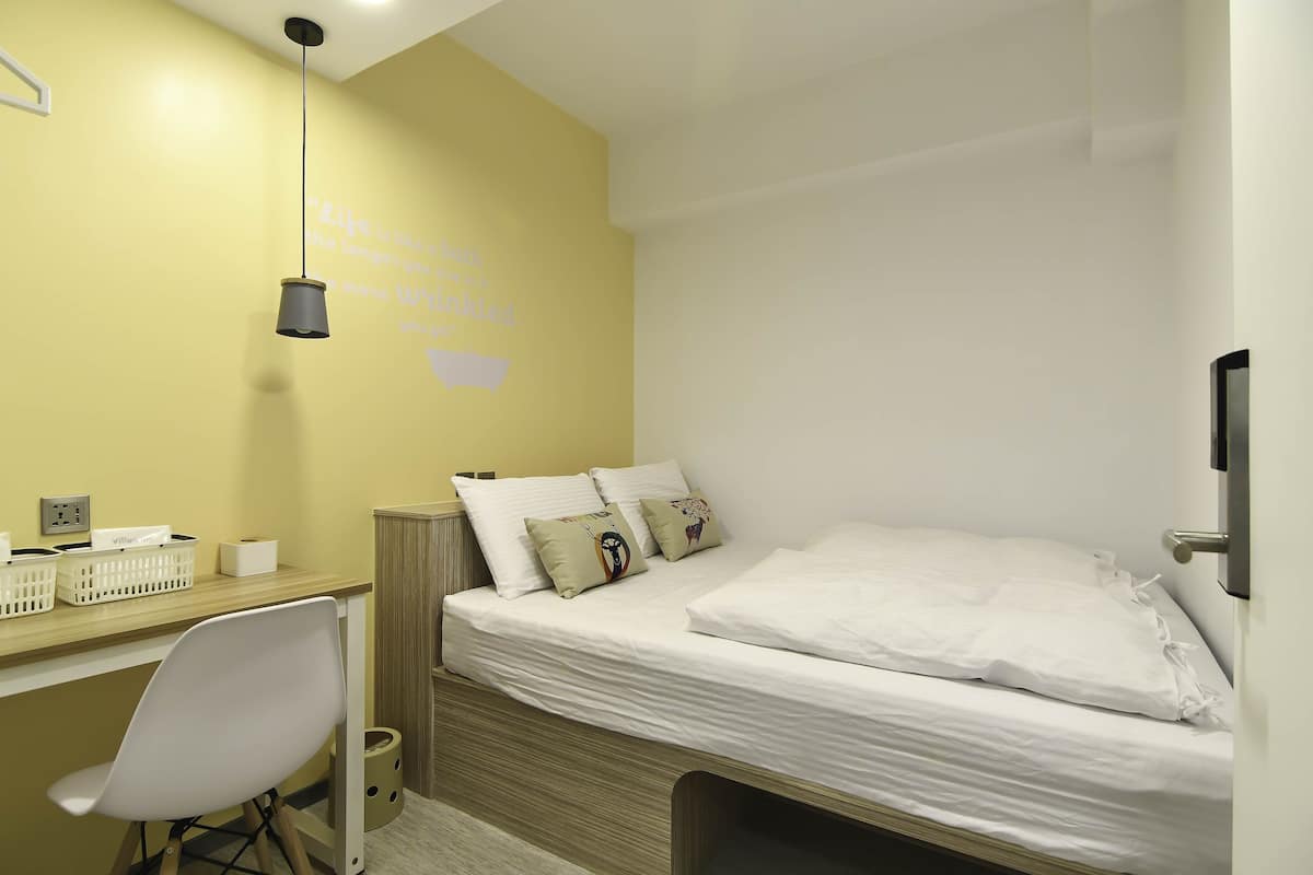 Ein Schlafzimmer mit Bett, Schreibtisch und Stuhl. An der Wand befindet sich ein Regal mit Körben und eine kleine Uhr.