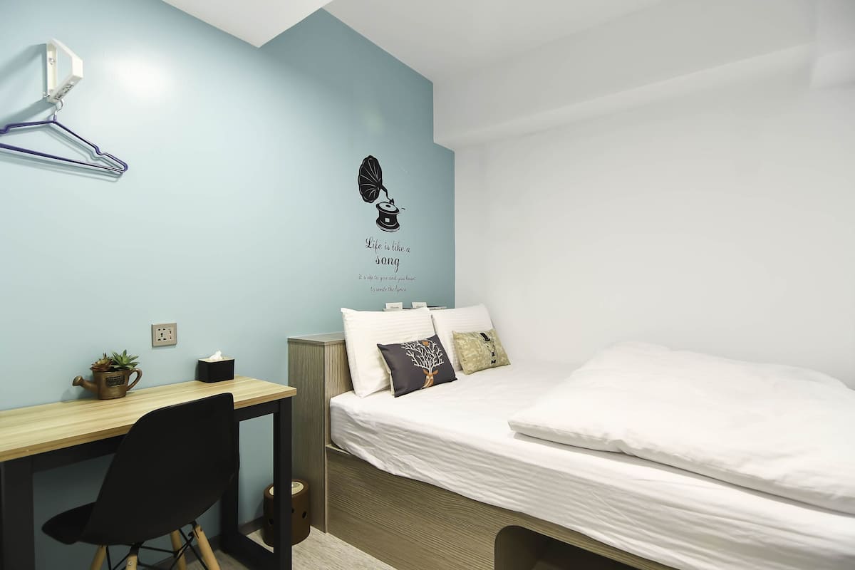 Ein Schlafzimmer mit einem Bett, einem Schreibtisch und einem Stuhl. An der Wand ist ein Heizlüfter angebracht, auf dem Schreibtisch steht eine kleine Pflanze.