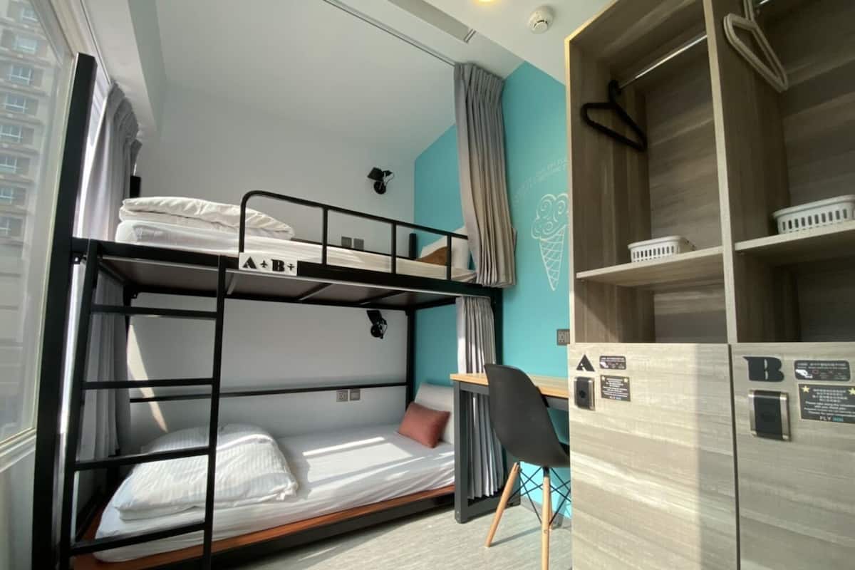 Ein Schlafraum mit Etagenbetten, Schreibtisch und Stuhl, einem kleinen Regal mit Gegenständen und einem Fenster mit Vorhängen.