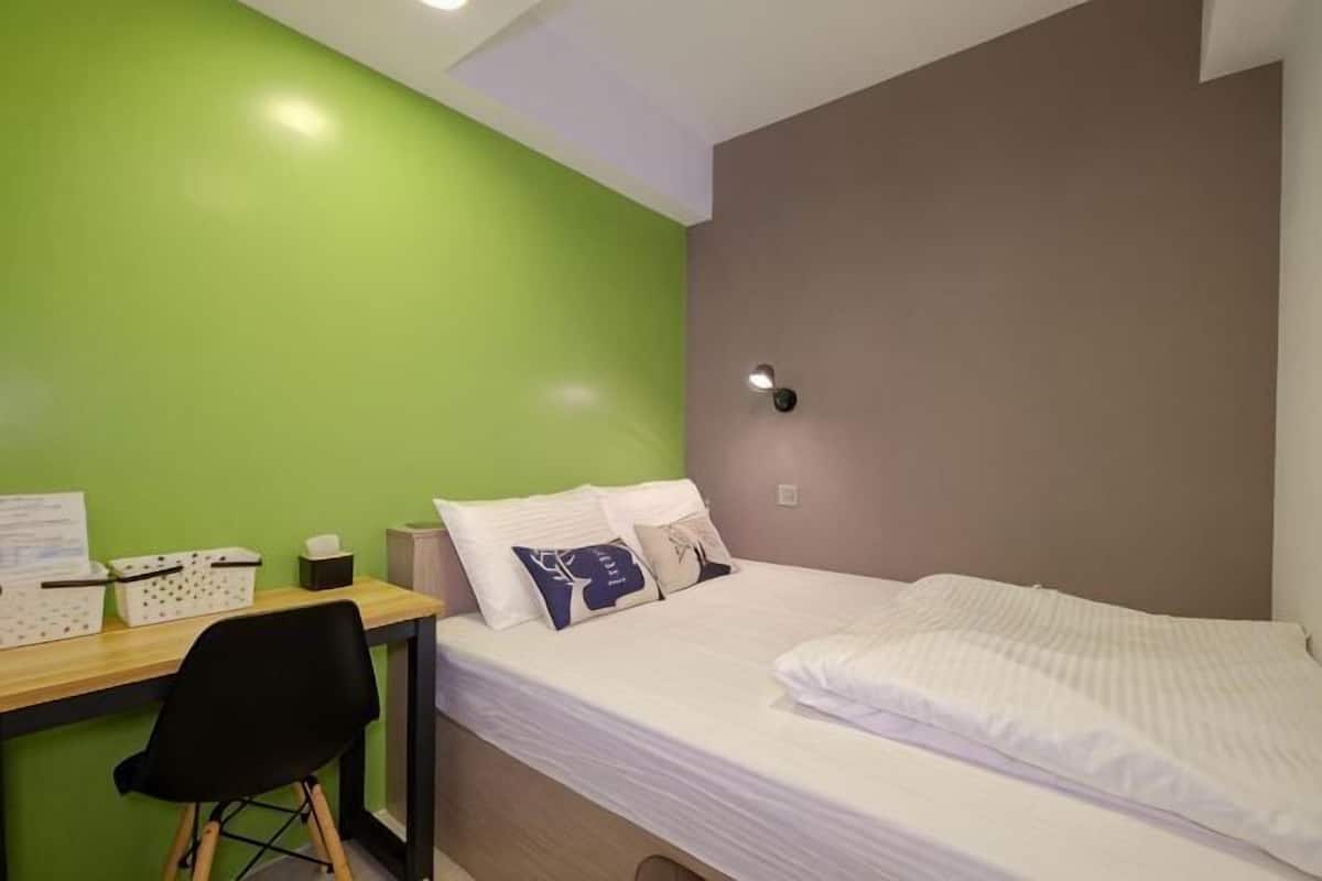 Ein Einzelbett mit weißer Bettwäsche, ein Schreibtisch mit Stuhl und eine grüne Wand.