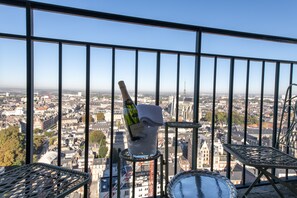 A balcony with a bottle of champagne and a glass of ice, overlooking a cityscape with buildings and a tower.