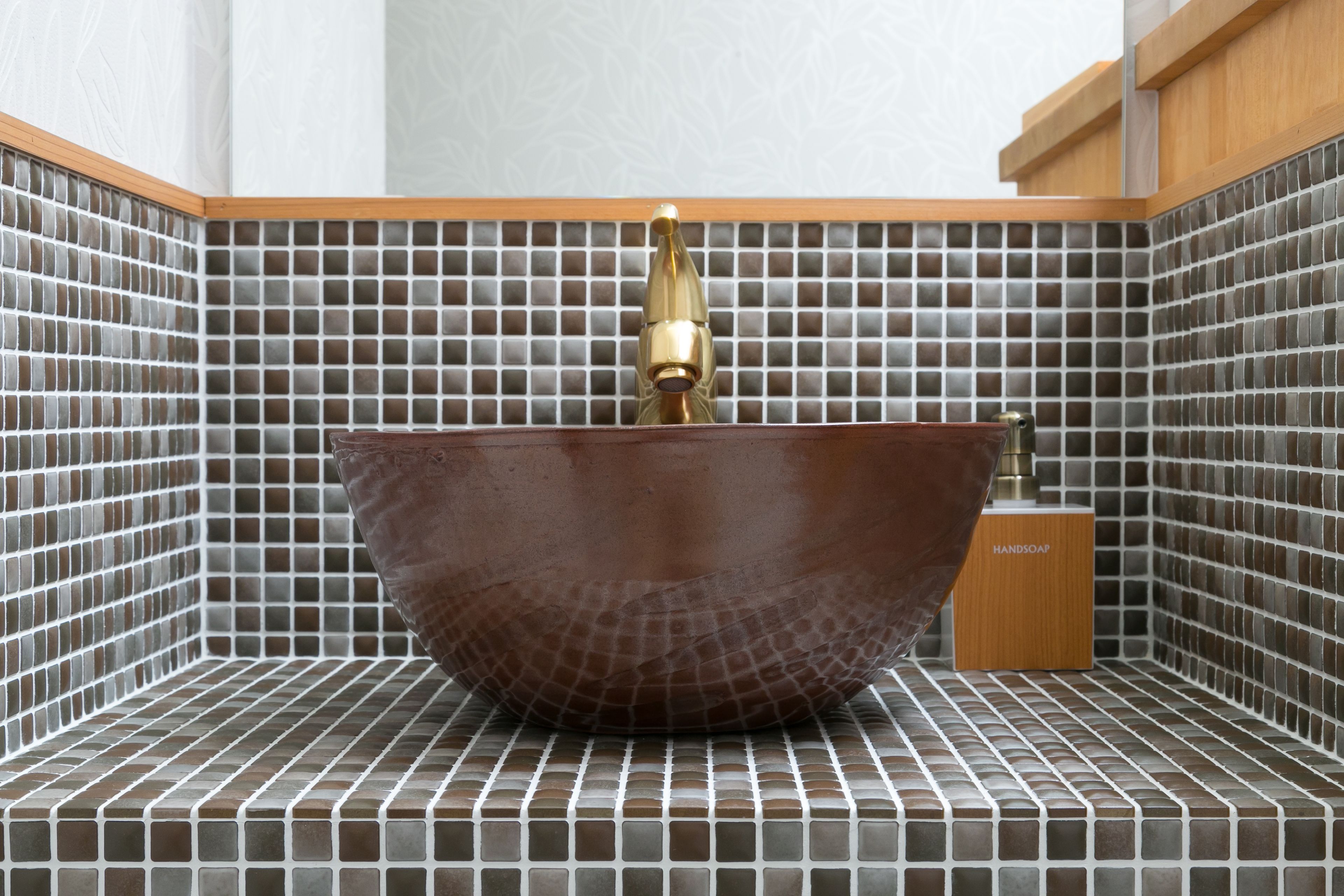 A bathroom with a large, round, brown ceramic sink with a gold faucet, set against a tiled wall with a grid pattern.