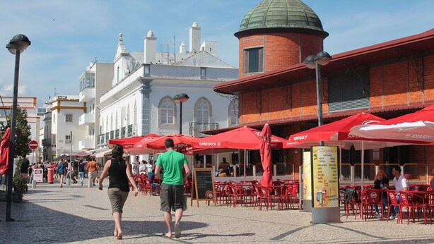 A cobblestone street with outdoor seating and red umbrellas, a brick building with a dome, and white buildings in the background. - Mar i Terra, the home of your vacation! (olhao)