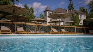 A swimming pool with lounge chairs and umbrellas, surrounded by a wooden fence and a large building with a tower in the background.
