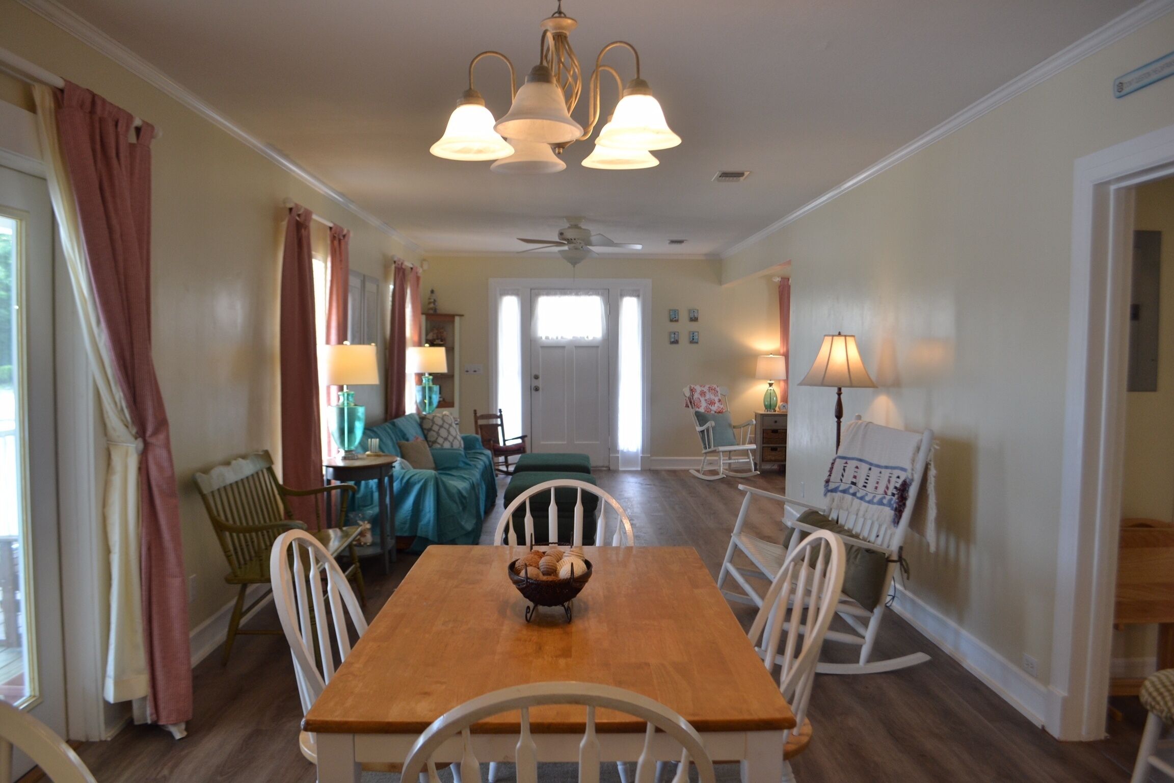 A dining area with a wooden table and white chairs, a ceiling fan, and a chandelier.