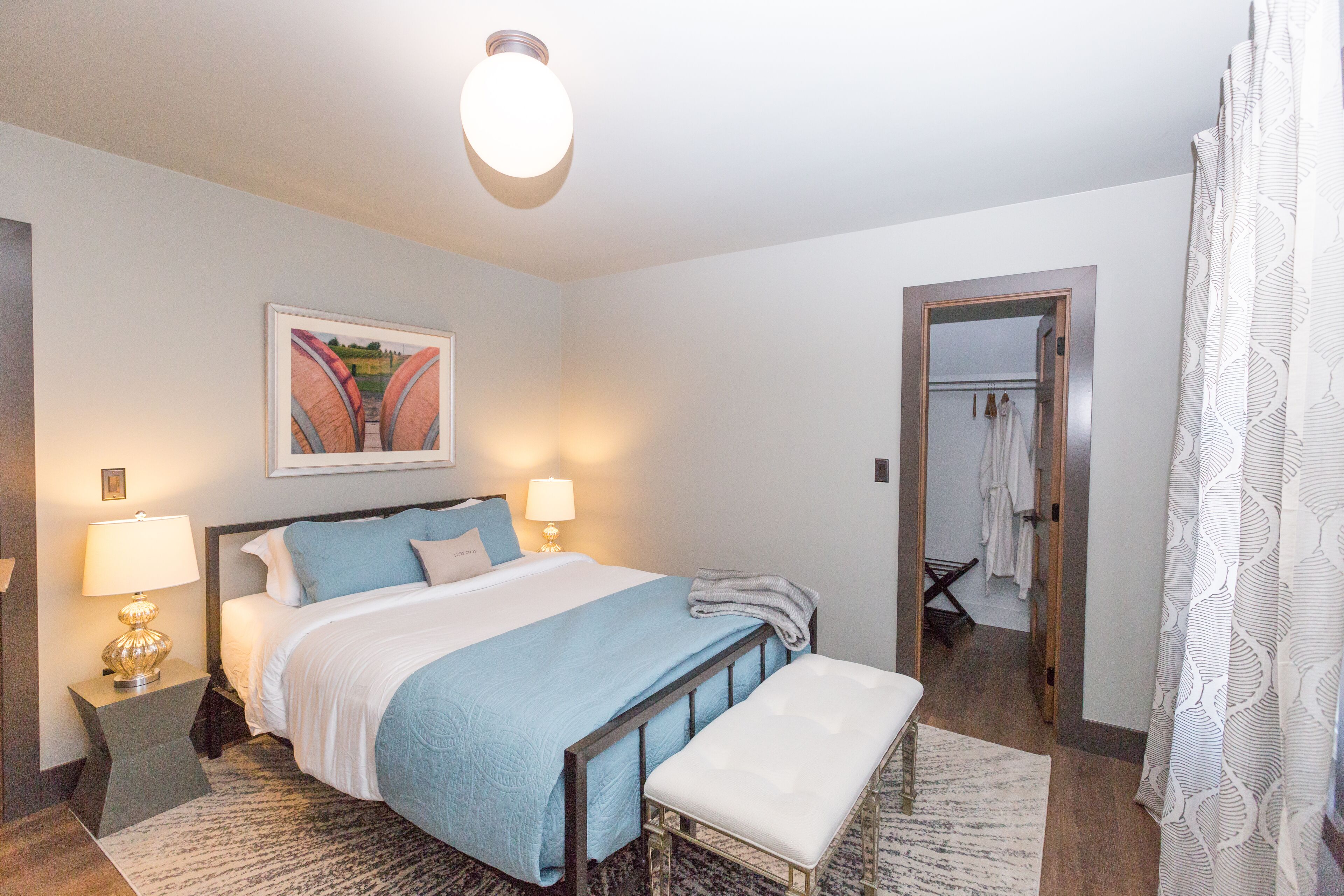 A bedroom with a bed, bedside tables, a nightstand, a bench, a wall-mounted lamp, a ceiling light, a door, and a mirror.