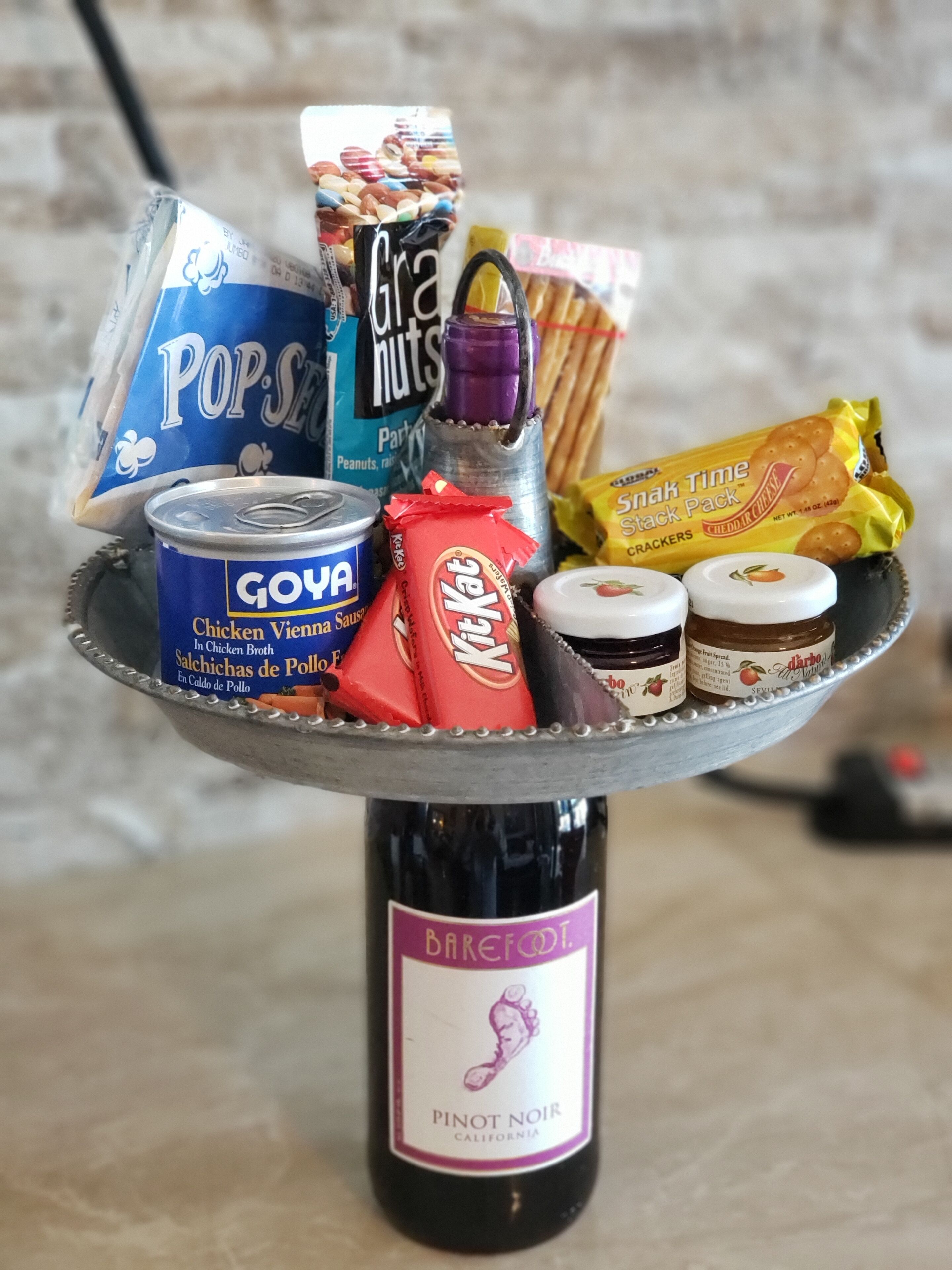 Verschiedene Snacks und Getränke auf einer Metalltablett, darunter eine Flasche Barefoot Pinot Noir, eine Dose Goya Geflügelwurst, eine Tüte Pop Secret, eine Packung Grahams, eine Flasche Schokolade und eine Tüte Snak Time Cracker.