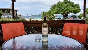 Mesa con botella de vino y una copa, con vista a una playa con gente y un árbol.