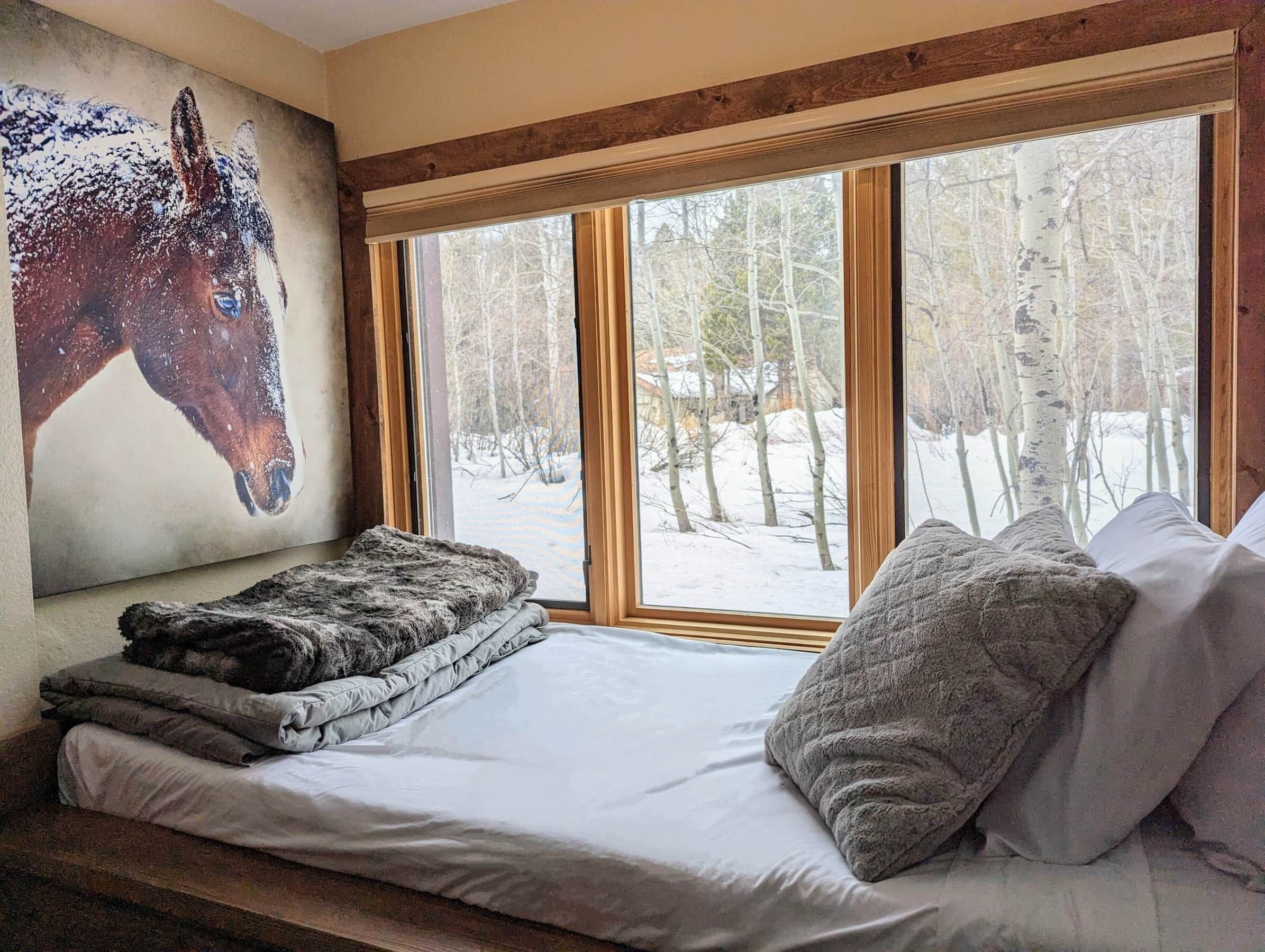 A cozy room with a large window, a bed with pillows, and a horse painting on the wall.