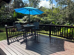 Une terrasse en bois avec une table et des chaises, un parasol bleu et, en arrière-plan, une végétation luxuriante.