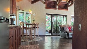 A spacious living area with a high ceiling, wooden beams, a dining table with chairs, a sofa, and a large window with a view of greenery. - Private Cottage In The Rain Forest TA065882726401 (Kailua-Kona)