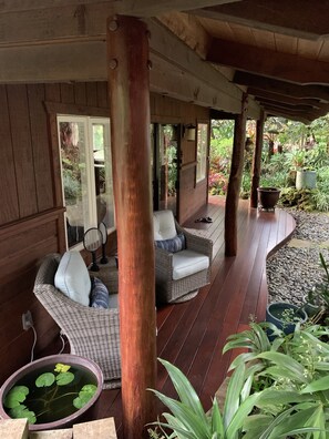 Een overdekt terras met rieten meubels, een houten vloer en potplanten.