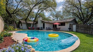 Un patio trasero con piscina, dos flotadores inflables, un porche con mobiliario exterior y una valla de madera.