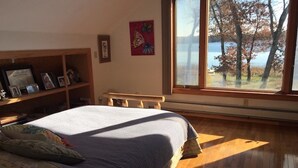 A bedroom with a large bed, wooden floor, a window with a view of trees and water, a bookshelf, and a framed picture on the wall.