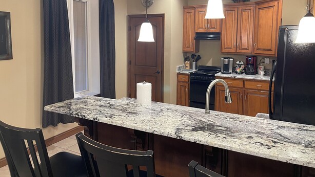 A kitchen with wooden cabinets, a granite countertop, a black refrigerator, and a window with dark curtains.