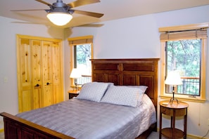 Quarto com cama, guarda-roupa de madeira, duas luminárias de cabeceira, ventilador de teto e janelas com persianas.