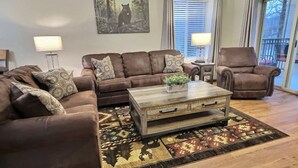 A living room with a brown leather sectional sofa, a wooden coffee table, and a patterned rug. There is a painting of a bear on the wall and a window with blinds. - Beautiful Downtown Gatlinburg Smokey mountain condo  (Gatlinburg)