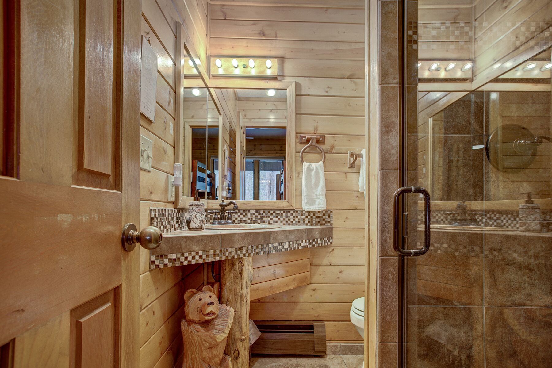 Ein Badezimmer in einer Holzhütte mit Holztür, Spiegel, Waschbecken mit kariertem Fliesenspiegel, Handtuchhalter und einer Duschkabine aus Glas.
