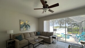 Una sala de estar con un sofá modular, una mesa de centro, un ventilador de techo y un ventanal con vistas a una zona de piscina.