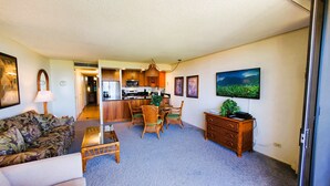 Ein Wohnzimmer mit Sofa, einem Esstisch mit Stühlen, einer Küchenzeile und einem Fernseher an der Wand.