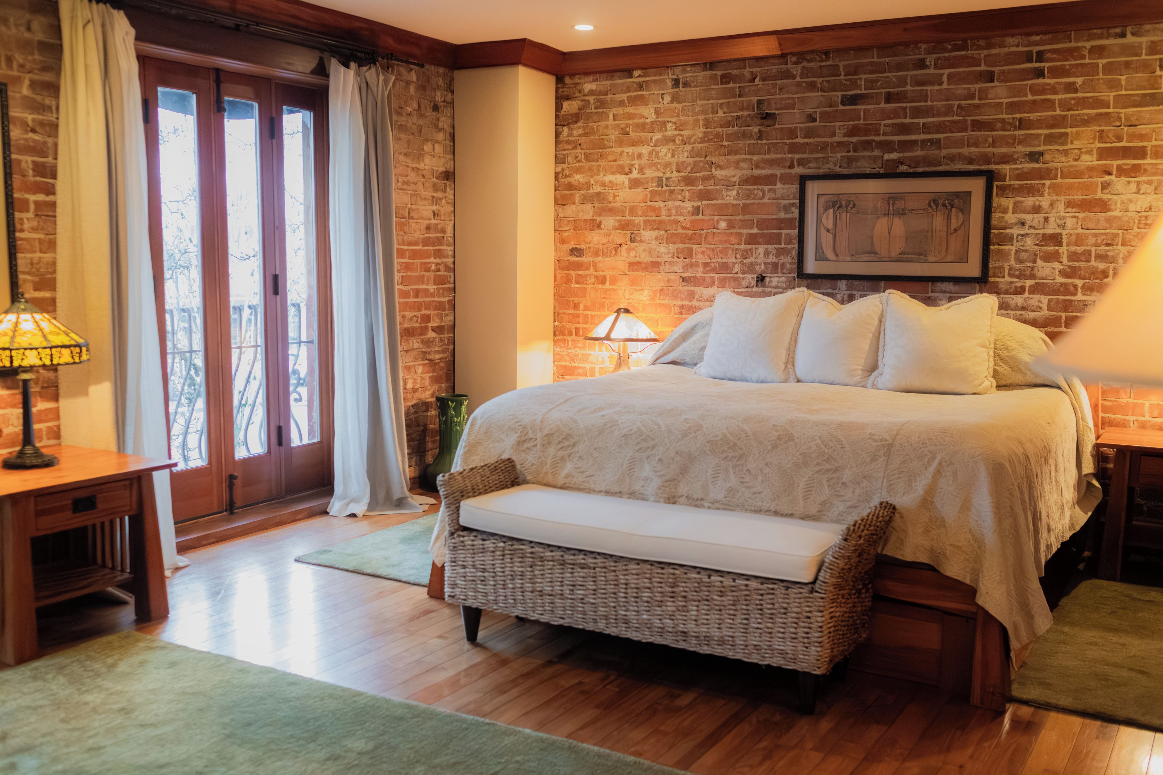 Une chambre avec un mur en briques, un lit avec une literie blanche, un banc en osier, une table de chevet avec une lampe et une fenêtre avec des rideaux.