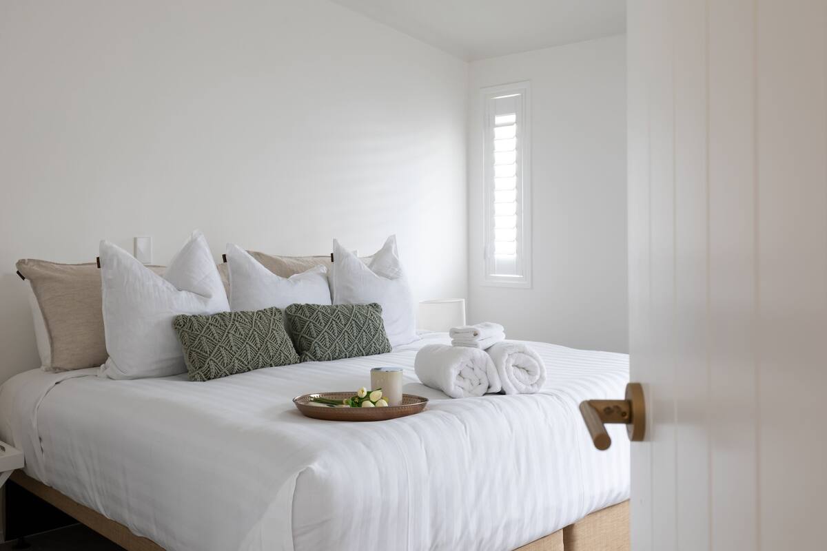 A neatly made bed with white linens, a tray with fruits and a drink, and a window with blinds.