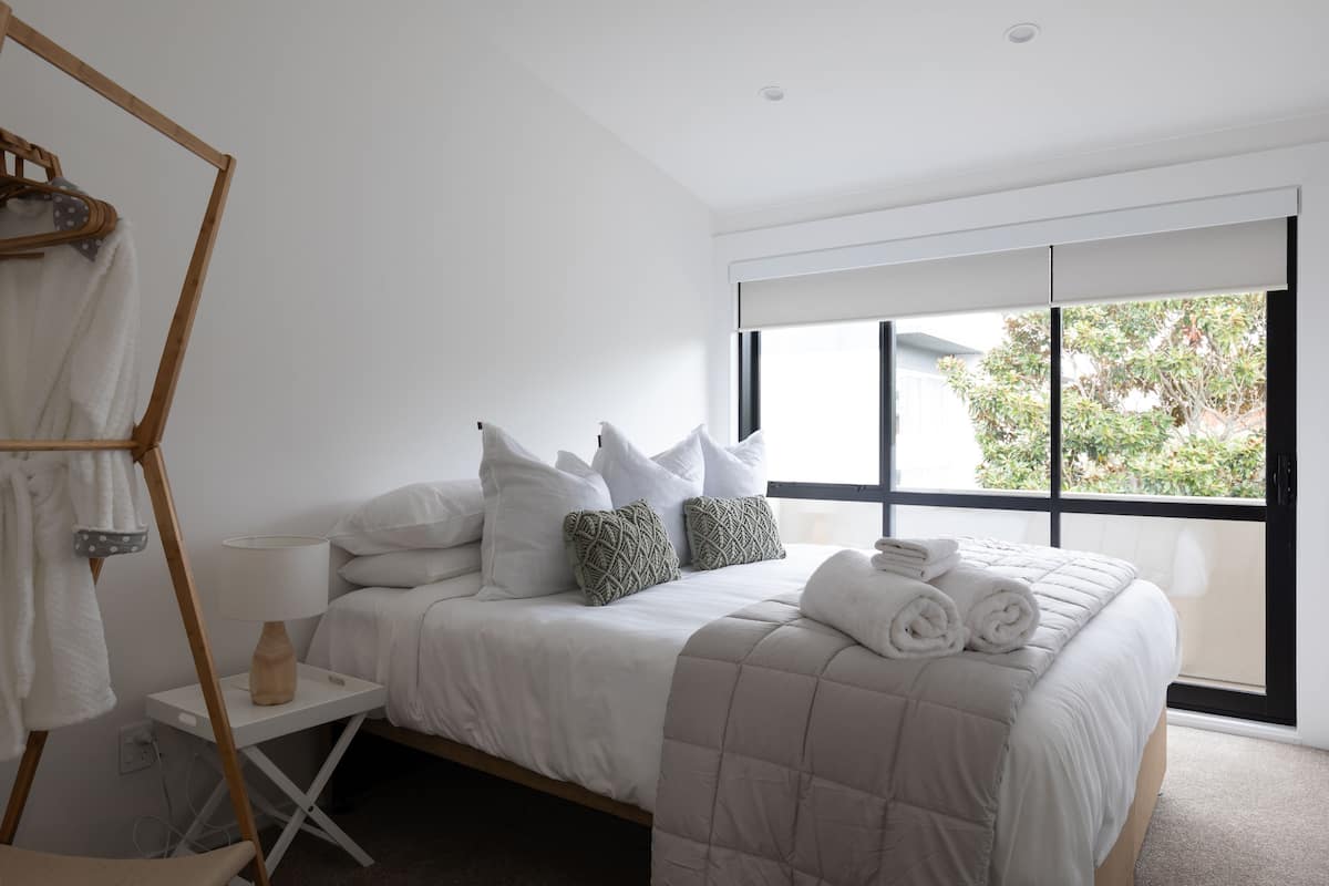A modern bedroom with a bed, pillows, a bedside table, a lamp, and a window with a view of greenery.