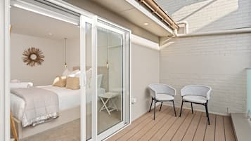 Un dormitorio con cama, una mesita y dos sillas. Hay una puerta corrediza de vidrio que da a un balcón con cubierta de madera.