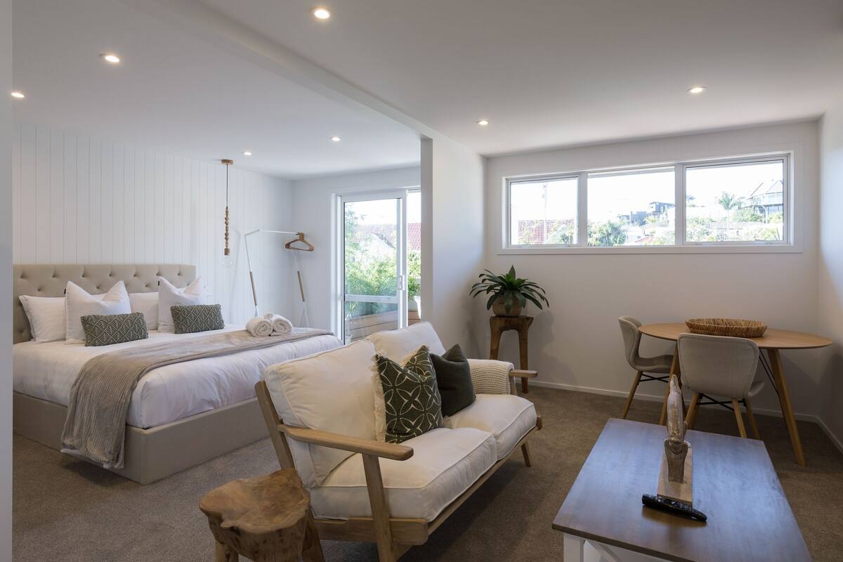 A modern bedroom with a large bed, a sofa, a dining table, and a potted plant.