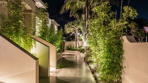 A nighttime view of a modern building with a well-lit pathway, surrounded by lush greenery and palm trees. - Stay Mission Bay (Auckland)