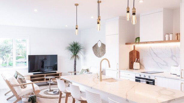 A modern kitchen with a large island, white cabinetry, and a built-in oven. There is a dining area with white chairs and a TV in the background. - Stay Mission Bay (Auckland)