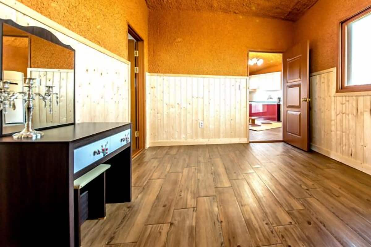 A room with wooden flooring, a wooden vanity with a mirror, and a window with a view of another room.