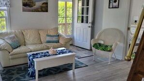Uma sala de estar com um sofá bege, uma mesa de centro branca, uma cadeira de vime e uma porta que dá para o exterior.