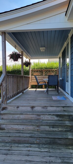 A covered wooden deck with a table and chairs, surrounded by a wooden fence and greenery.