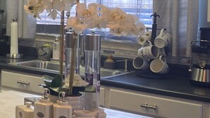 Une cuisine avec une orchidée blanche dans un vase en verre, une machine à café et une pile de tasses blanches sur le comptoir.
