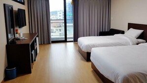 A hotel room with two beds, a desk, a TV, and a view of the city through large windows. - Chilsimni Hotel (Seogwipo)