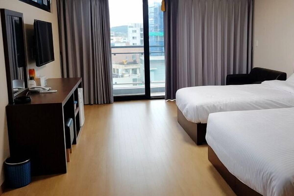 A hotel room with two beds, a desk, a TV, and a view of the city through large windows.