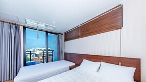 A hotel room with a large bed, a smaller bed, a balcony with curtains, and a view of the city. - Chilsimni Hotel (Seogwipo)