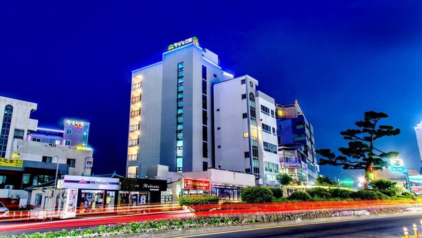 A multi-story hotel with illuminated signage, a parking area, and a flower-lined street. - Chilsimni Hotel (Seogwipo)