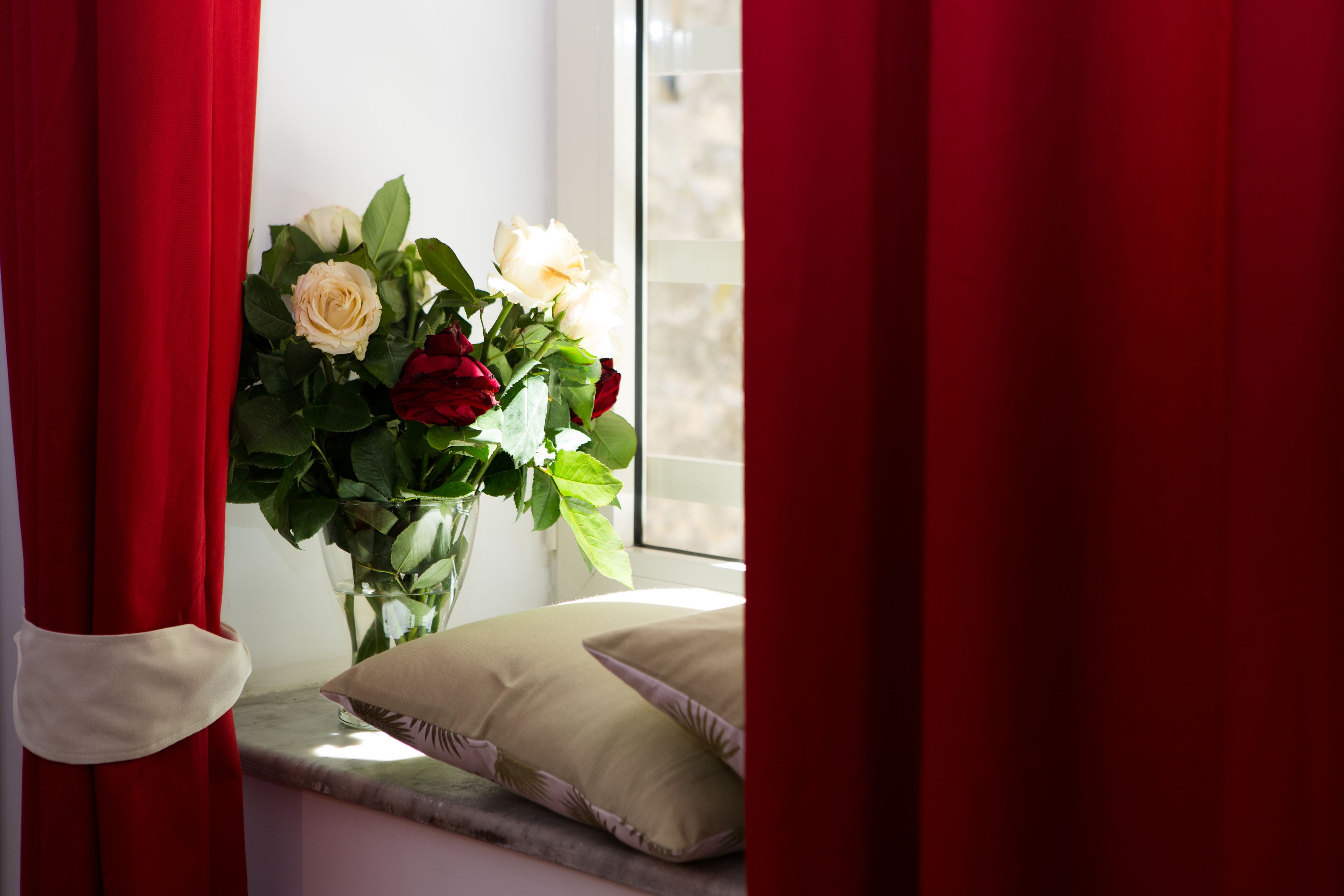 A window with red curtains, a vase with white and red roses, and a cushion on a ledge.