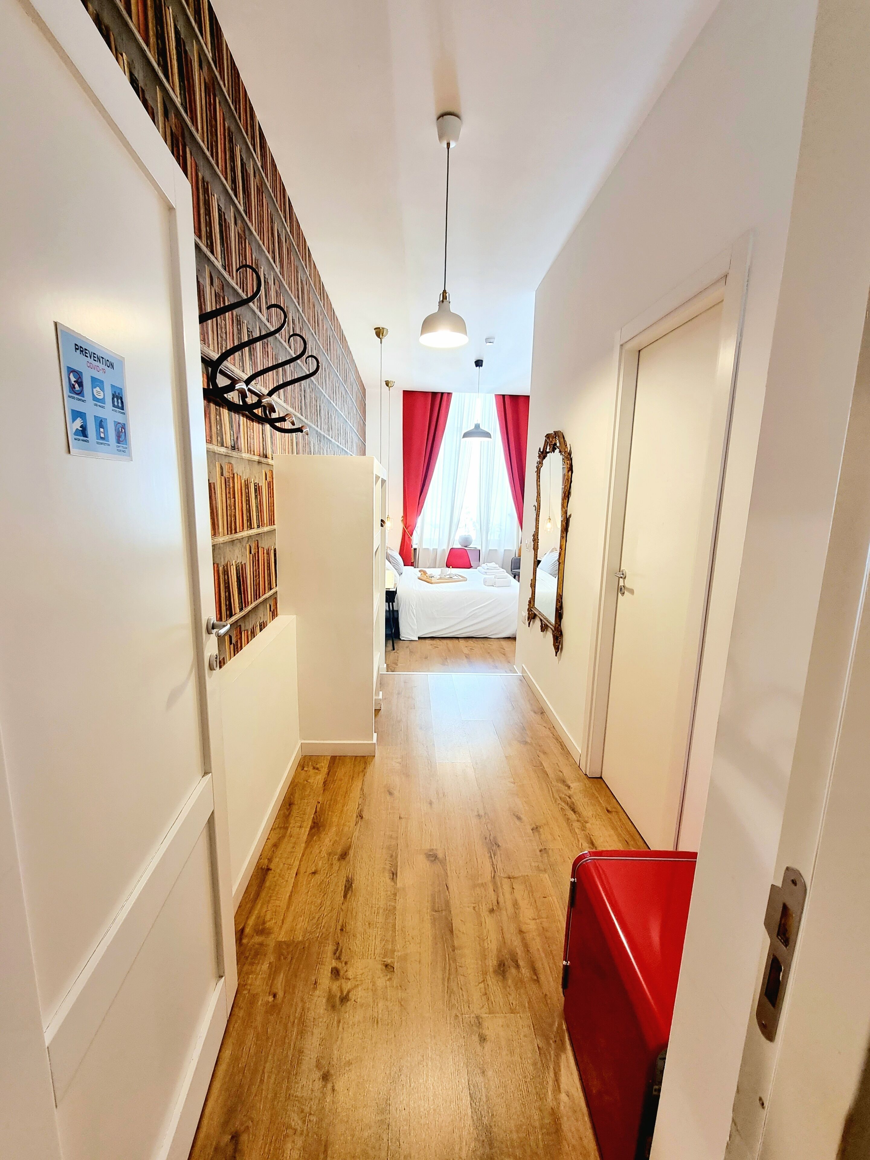 A hallway with a wooden floor, a red chair, a bed with white bedding, and a bookshelf.
