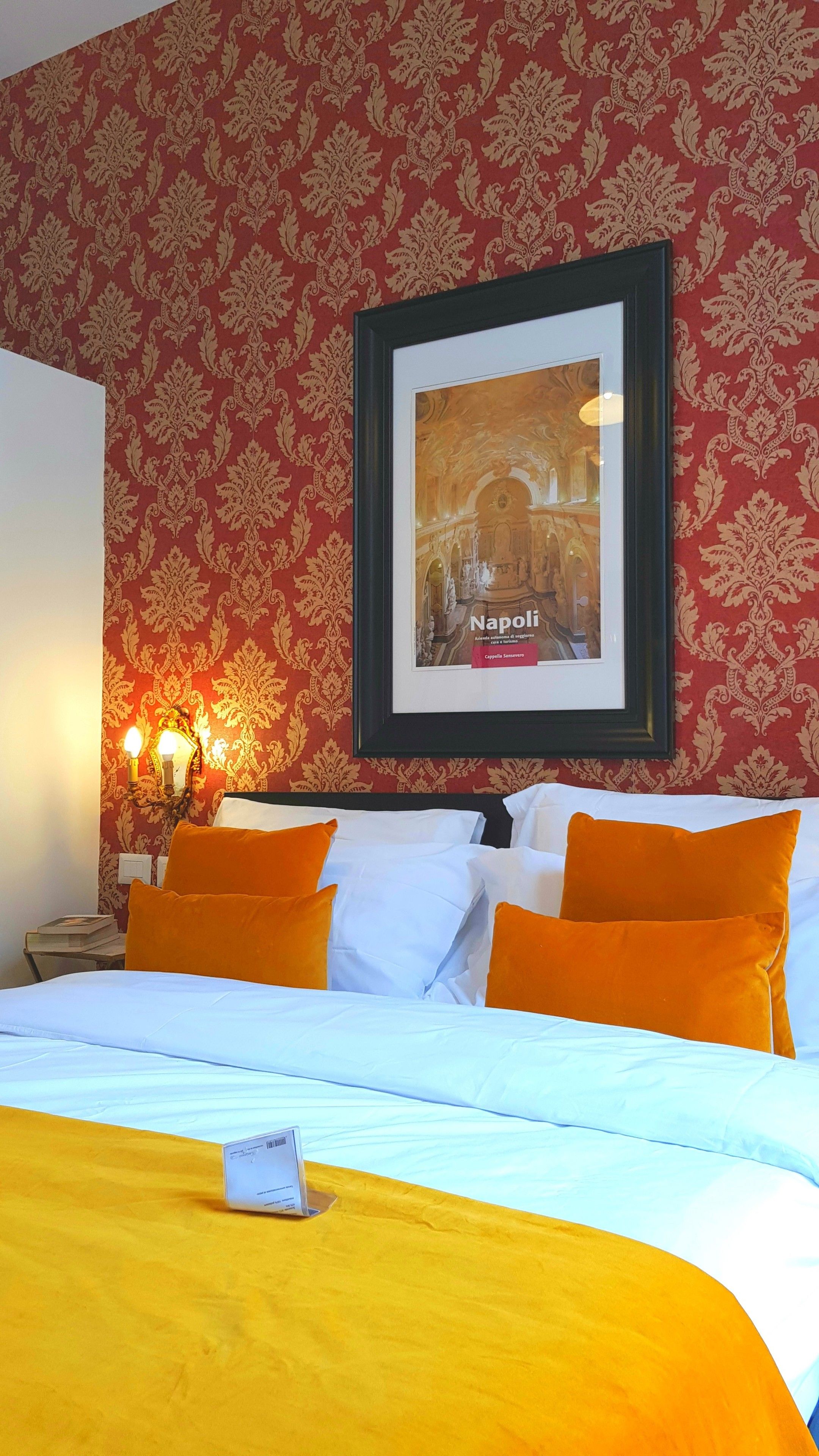 Una habitación de hotel con un cuadro enmarcado de un edificio histórico, una cama con ropa de cama blanca y naranja, y una mesita de noche con una lámpara.