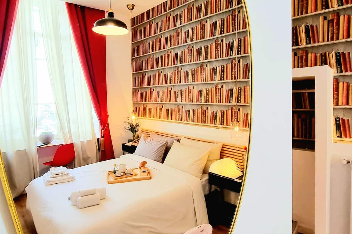 Una habitación de hotel con una cama grande, una mesita de noche, un espejo, una estantería y una silla roja.