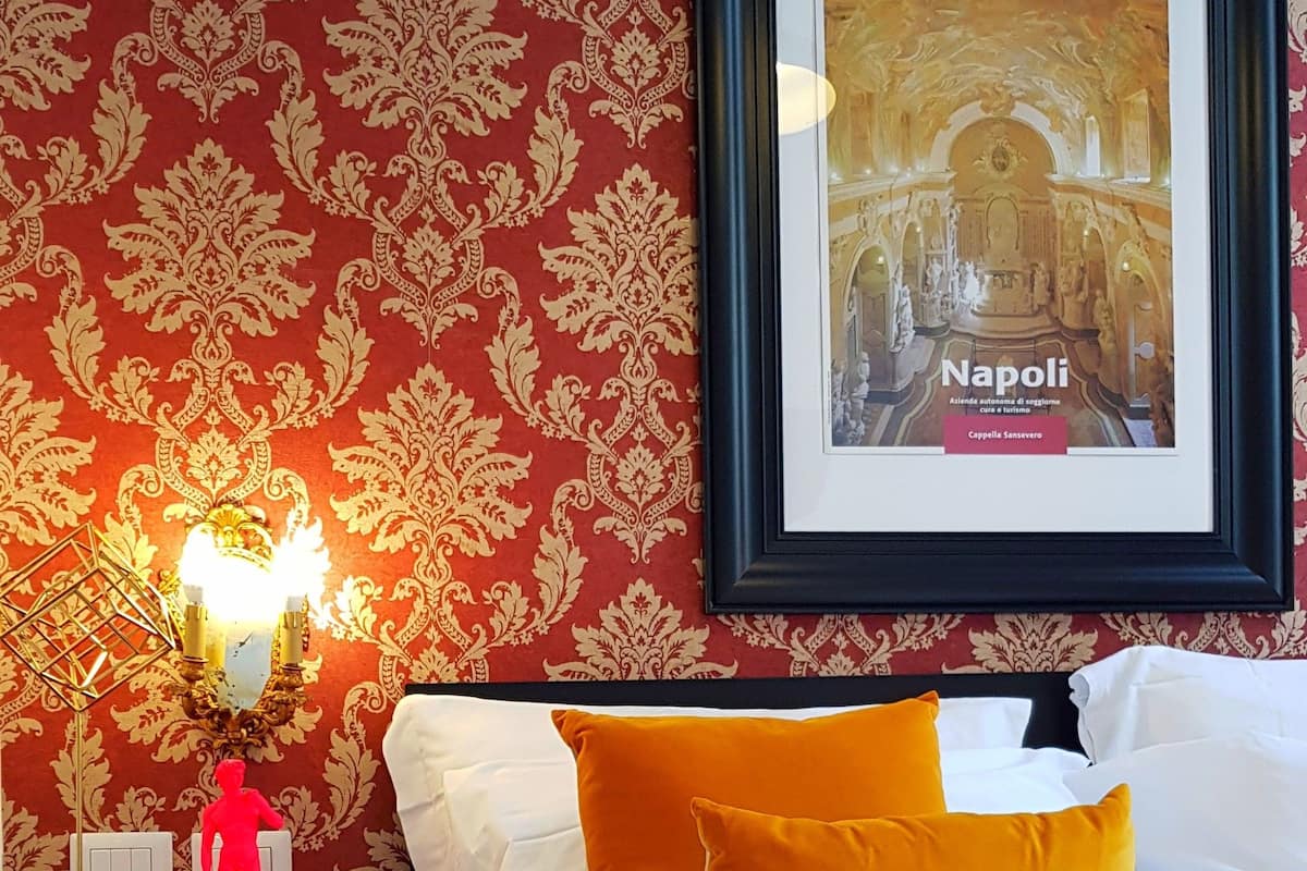 Habitación de hotel con una cama, un cuadro enmarco de un edificio histórico, una mesita de noche con una lámpara y una estatua roja.