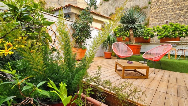 Una terraza de madera con una mesa y dos sillas rojas, rodeada de plantas en macetas y un muro de piedra.