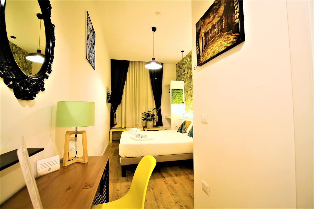 Una habitación de hotel con cama, escritorio con lámpara, silla, espejo y un cuadro en la pared.